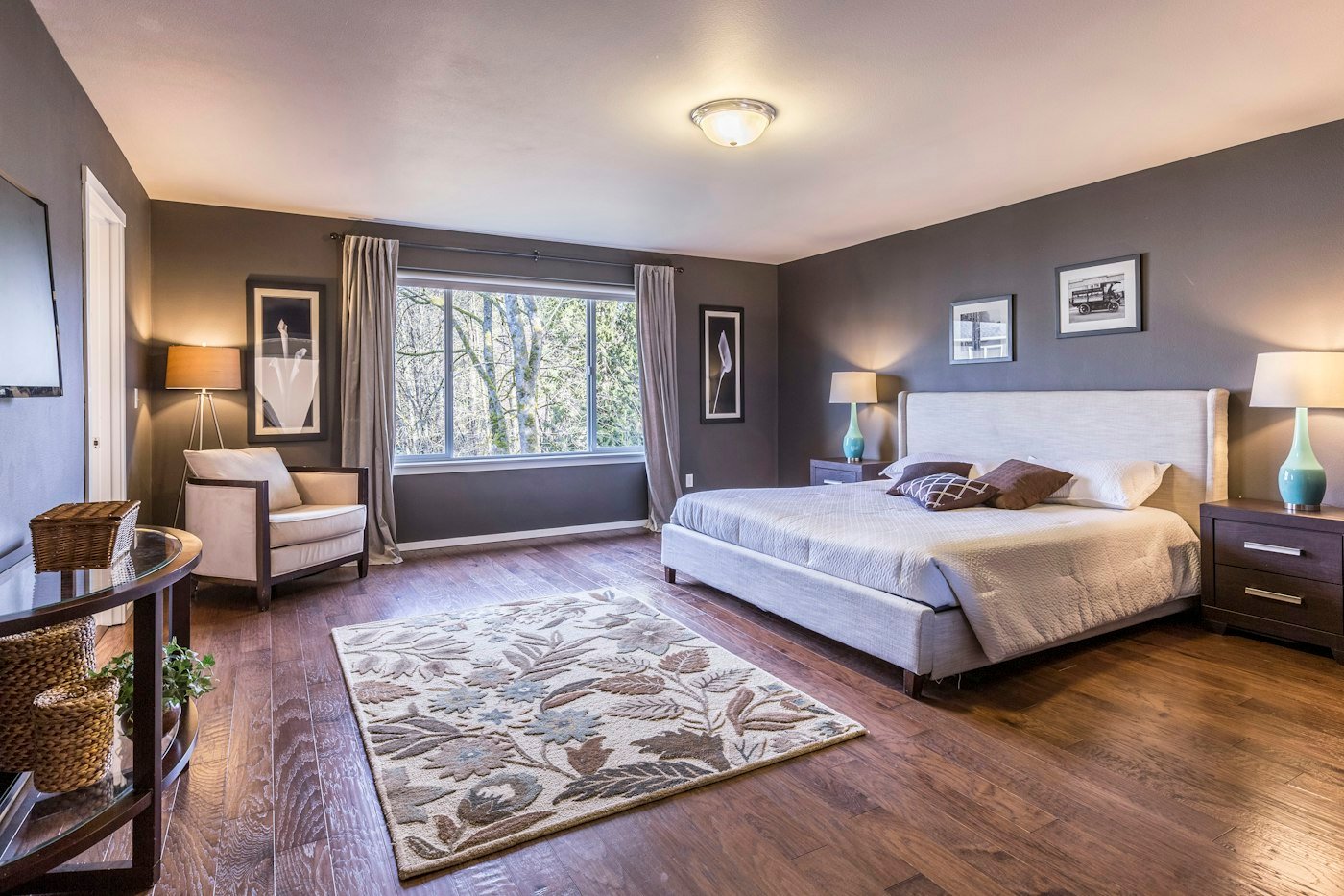 A pale bed against a grey wall, a floral rug, and trees visible through the window